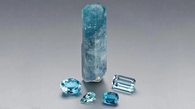 Cristallo di acquamarina grezzo affiancato da acquamarine tagliate di diverse forme